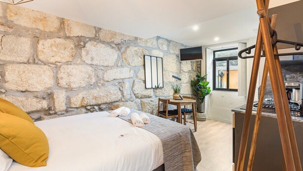 Une chambre avec un mur en pierre, un lit avec des serviettes roulées, une chaise en bois, une petite table avec une plante, et un poêle en arrière-plan.