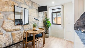 Une pièce avec un mur en pierre, une table à manger en bois, des chaises, un téléviseur fixé au mur et une fenêtre donnant sur l’extérieur.