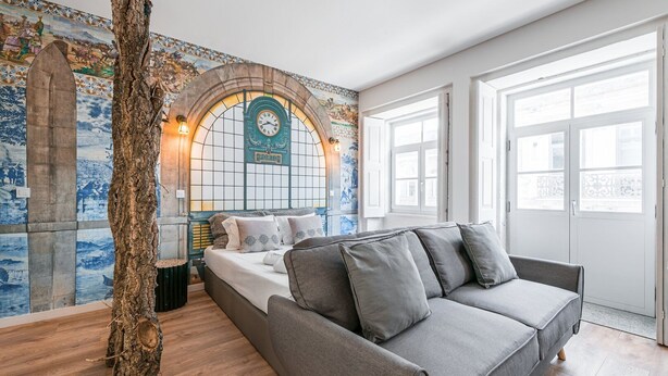 Een slaapkamer met een bed, een grijze bank, een groot raam en een muur met een klok en kunstwerken.