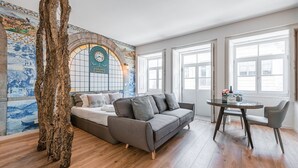 Een moderne woonkamer met een grijze bank, een klein rond tafeltje met een fles en glazen, en een groot raam met witte kaders.