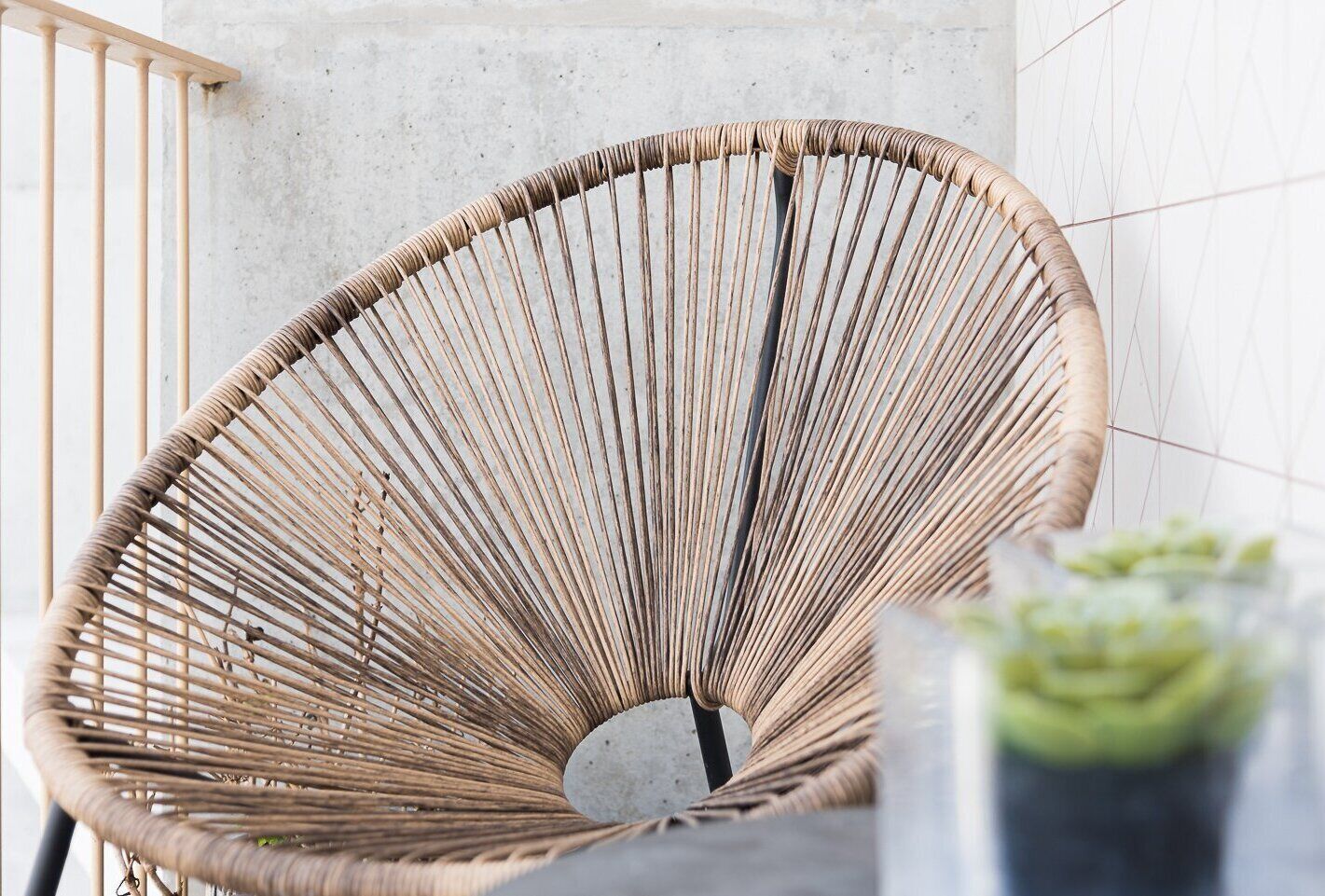 A wicker chair with a curved backrest, positioned against a concrete wall with a geometric tile pattern.