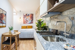 Quarto de hotel compacto com uma cama, uma pequena mesa de jantar, área de cozinha com pia e fogão, e uma parede de pedra.