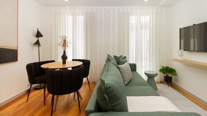 Una sala de estar moderna con una mesa redonda de madera, sillas negras, un sofá verde, una mesita auxiliar con una planta y un televisor montado.