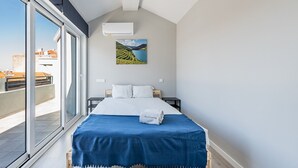 A modern bedroom with a large bed, a view of the city through a sliding glass door, and a painting of a landscape on the wall.