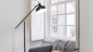 Ein Zimmer mit Fenster, einer schwarzen Stehlampe, einem grauen Sofa und einem Kissen mit Muster.