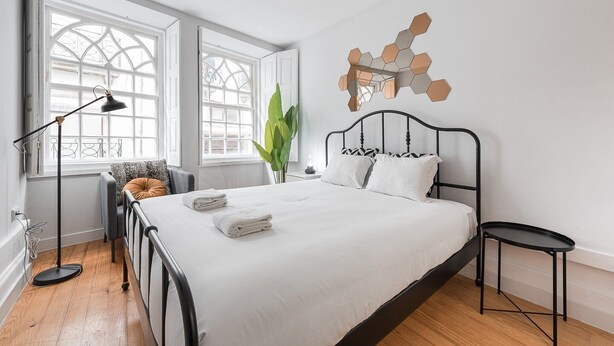 Ein modernes Schlafzimmer mit einem großen Bett, zwei Fenstern, einer Stehlampe, einem Sessel und einem kleinen Tisch.