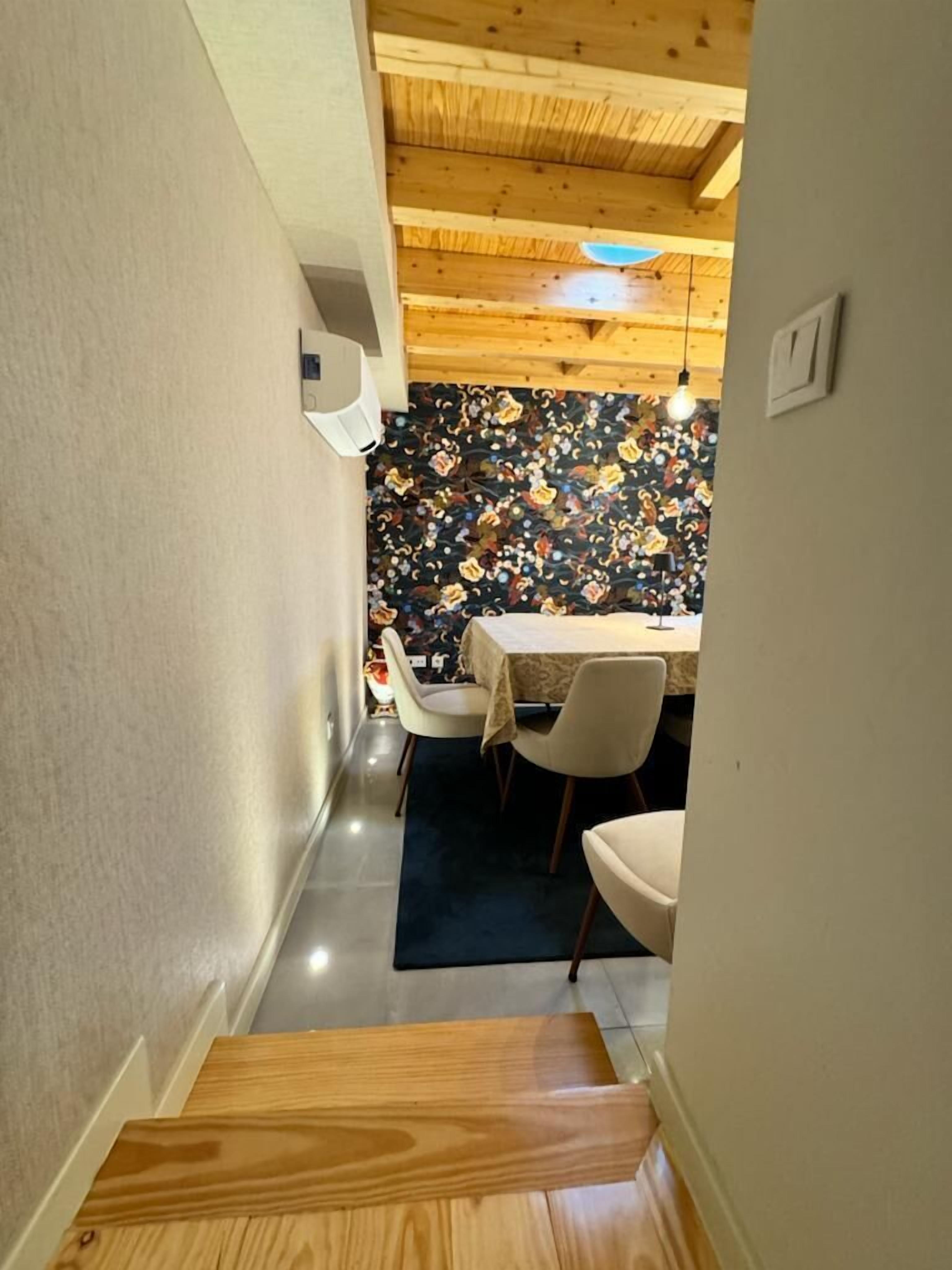A narrow hallway with a wooden staircase leading to a dining area. The dining area features a table with chairs and a colorful floral wallpaper.