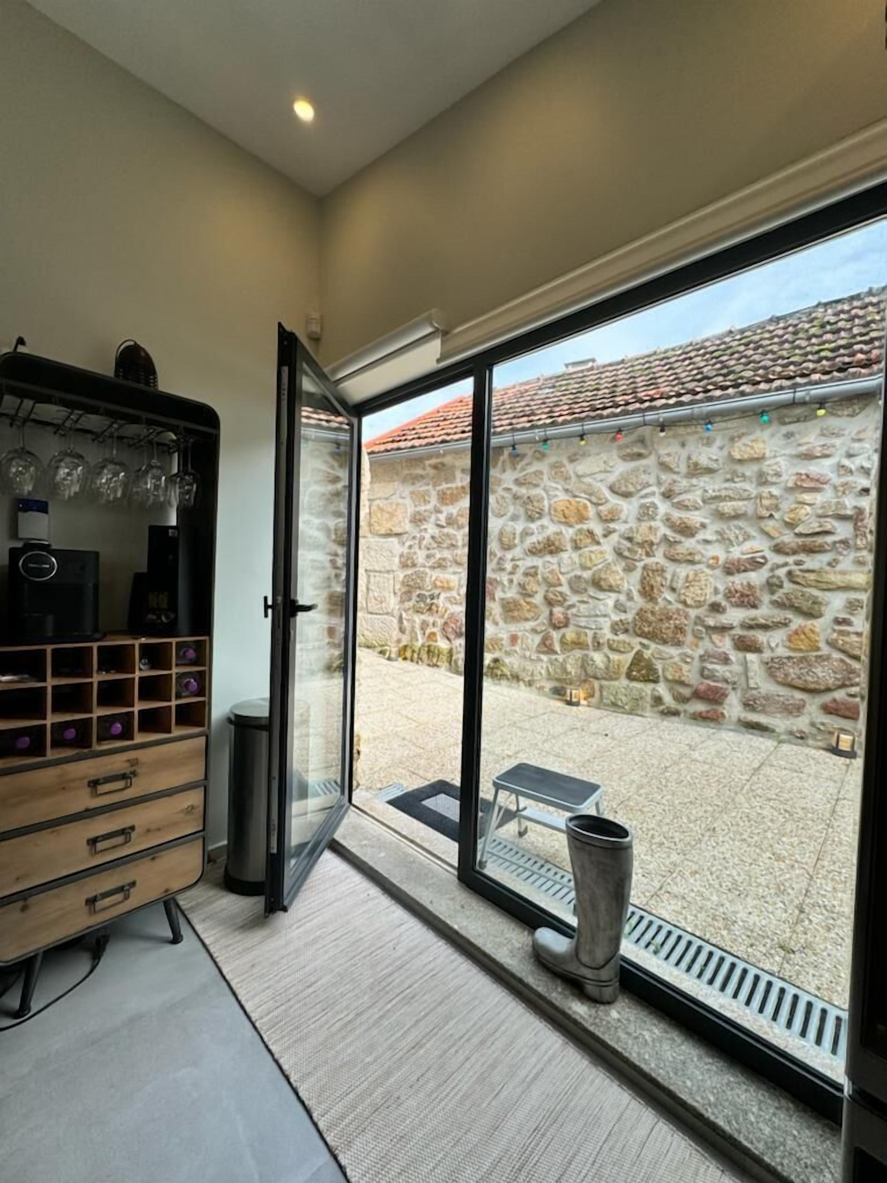 Une pièce avec un mur en pierre, un buffet en bois contenant des verres à vin et une porte-fenêtre coulissante donnant sur un espace extérieur doté d’un mur en pierre et d’un banc en métal.