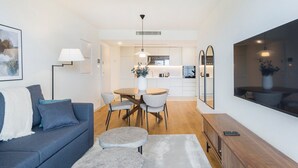 A modern living room with a sofa, a dining table, and chairs, a kitchen in the background, and a large TV on the wall. - GuestReady - Asaf's Magnolias Retreat (Vila Nova de Gaia)