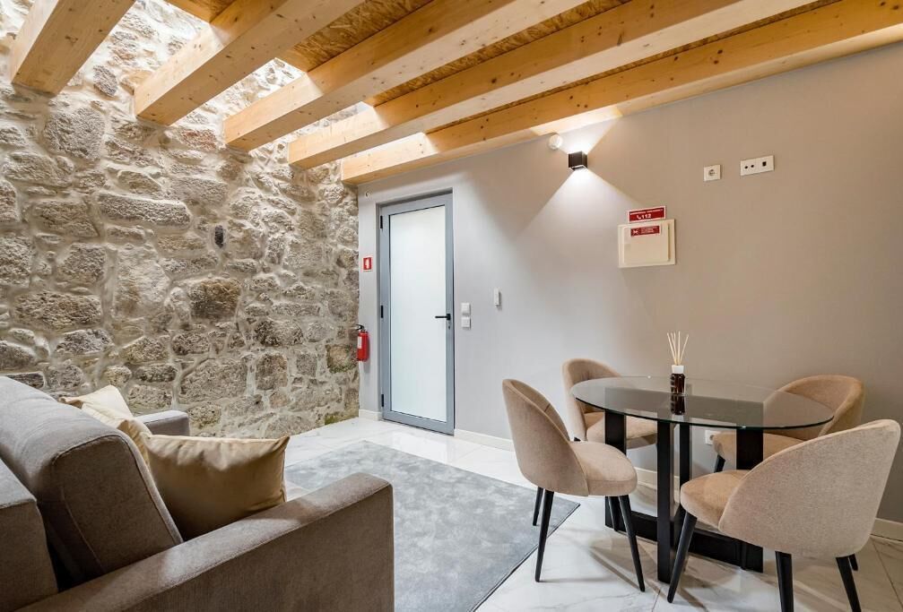 A cozy room with a stone wall, a wooden ceiling, a glass door, a round table with chairs, and a sofa.