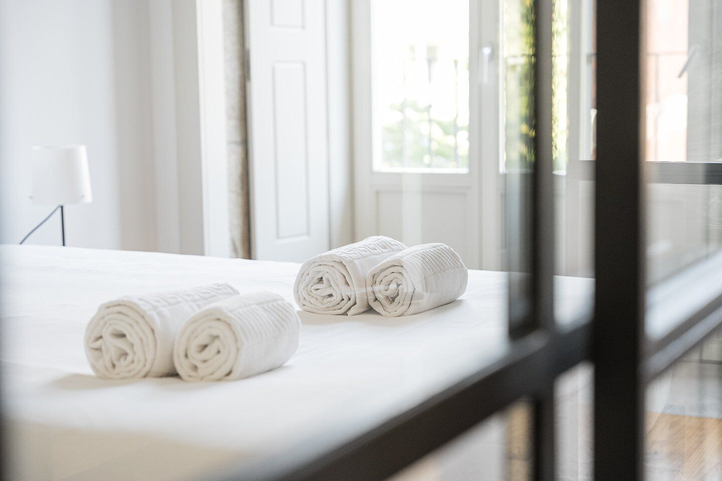 A neatly made bed with rolled white towels and a window with a view of greenery.
