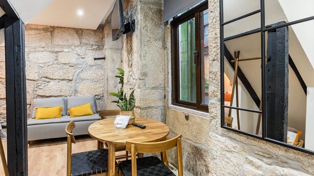 A cozy room with a stone wall, a wooden table with chairs, a sofa with yellow cushions, and a window with a view of the outside. - GuestReady - Caldeireiros Building (Porto)