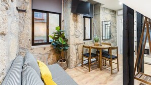 A room with a stone wall, a wooden table with chairs, a sofa with patterned cushions, and a television mounted on the wall. - GuestReady - Caldeireiros Building (Porto)