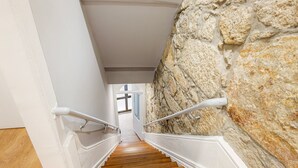 A staircase with wooden steps and white handrails, leading to a room with a stone wall. - GuestReady - Caldeireiros Building (Porto)