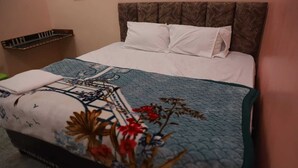 A neatly made bed with a patterned bedspread and pillows, a headboard, and a small shelf unit against the wall. - Shri Laddu Gopal Guest House (Mathura)