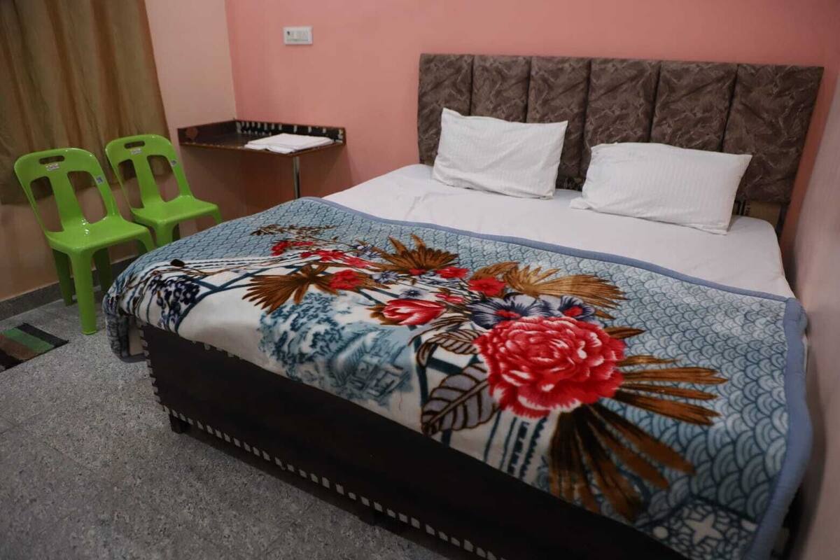 Ein Hotelzimmer mit einem Bett, zwei Kissen, einer blumigen Tagesdecke, zwei grünen Stühlen und einem kleinen Tisch mit einer gefalteten weißen Tischdecke.