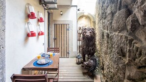 Ein schmaler Außenbereich mit Essplätzen, eine rustikale Steinwand, ein Holztisch gedeckt mit Tellern und Gläsern, sowie eine Löwenskulptur.