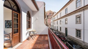 Une rue étroite, bordée de bâtiments des deux côtés, un balcon avec une table et des chaises, et des voitures garées.