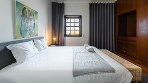 Une chambre d’hôtel avec un grand lit, une table de chevet avec une lampe, une fenêtre avec des rideaux, une armoire en bois et un tableau au mur.