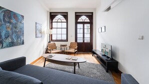 Un salon moderne avec un canapé, deux fauteuils, une table basse et un téléviseur. Il y a deux portes avec fenêtres, un tableau au mur et une lampe.