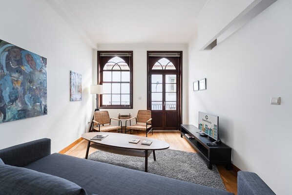 A modern living room with a sofa, two armchairs, a coffee table, and a TV. There are two doors with windows, a painting on the wall, and a lamp.