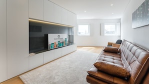 A modern living room with a brown leather sofa, a flat-screen TV mounted on a white cabinet, and a large window providing natural light. - GuestReady - Mompilher Apartments (Porto)