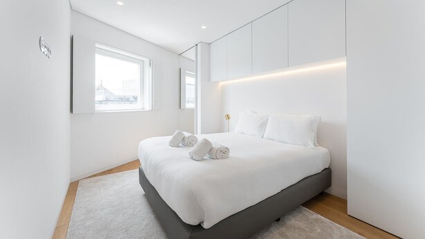 A modern bedroom with a large bed, white linens, a wooden floor, and a large window. - GuestReady - Mompilher Apartments (Porto)