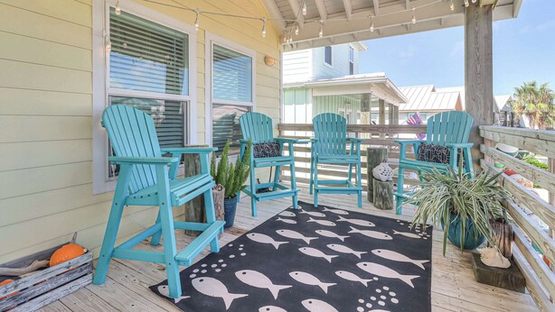 A covered porch with teal chairs, a black rug with fish patterns, and a wooden railing. - Brito Beach- View of Pond | Propane Grill (Port Aransas)