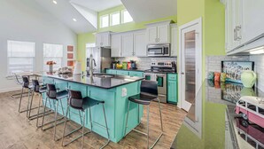 A modern kitchen with a central island, bar stools, and a dining area with a table and chairs. - Brito Beach- View of Pond | Propane Grill (Port Aransas)
