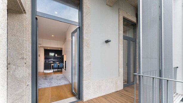 Un apartamento moderno con terraza de madera, puerta corrediza de vidrio que da a una sala de estar con sofá y mesa de centro, y un pequeño balcón con barandilla de metal.