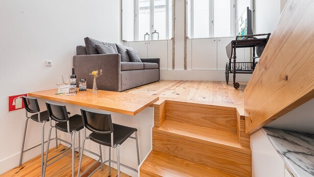Ein gemütlicher Wohnraum mit einer Couch, einem Esstisch mit Stühlen, einer kleinen Küchenzeile und Blick durch ein großes Fenster.
