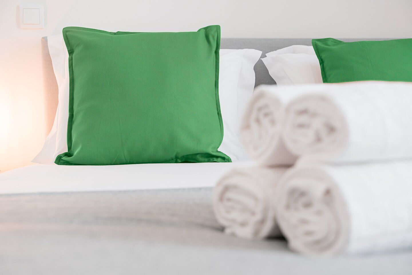 Een net opgemaakt bed met groene en witte kussens, en opgerolde witte handdoeken aan de zijkant.
