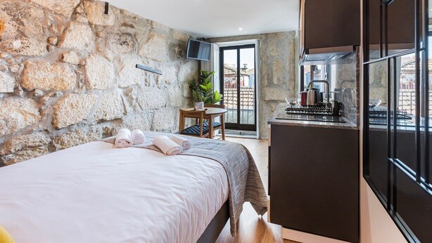 A bedroom with a stone wall, a bed with rolled towels, a small table with a plant, and a kitchen area with a stove and cabinets. - GuestReady - Caldeireiros Building (Porto)