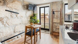 A compact kitchen with a stone wall, a dining area with a table and chairs, a TV mounted on the wall, and a view of the outside through a glass door. - GuestReady - Caldeireiros Building (Porto)