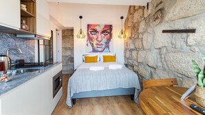 A compact hotel room with a bed, a small kitchenette, a stone wall, and a painting above the bed. - GuestReady - Caldeireiros Building (Porto)