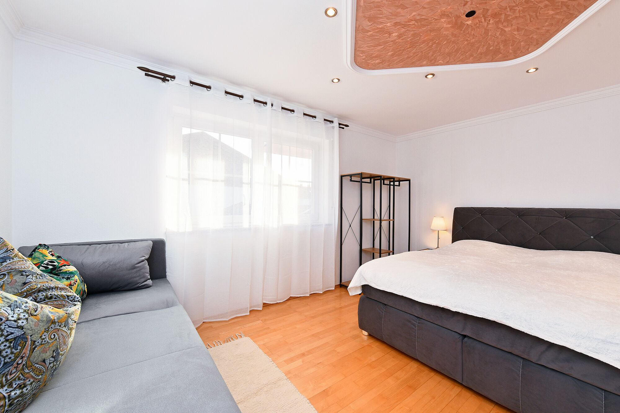 A modern bedroom with a grey sofa, a large bed, and a tall black shelving unit.