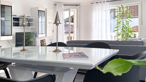 A modern living room with a glass dining table, a grey sofa, and a large window with a view of buildings.