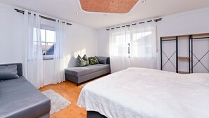 A modern bedroom with a bed, a grey sofa, a small seating area with cushions, and a wooden wardrobe.