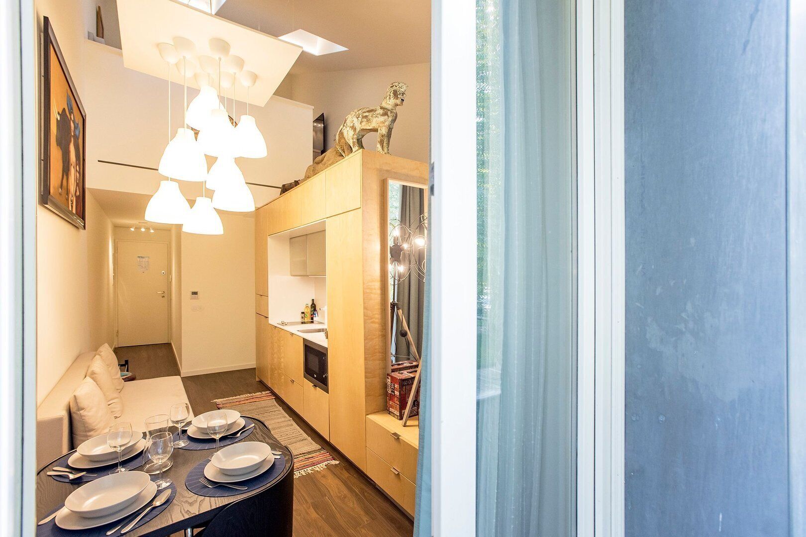 Una habitación de hotel moderna con comedor, sofá, pequeña cocina y un amplio ventanal con cortinas.