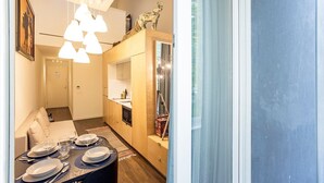 A modern hotel room with a dining area, a sofa, a small kitchen, and a large window with curtains.