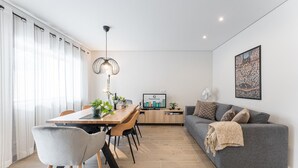 Un salon moderne avec un canapé gris, une table à manger avec des chaises, un luminaire suspendu et un téléviseur posé sur un meuble en bois.
