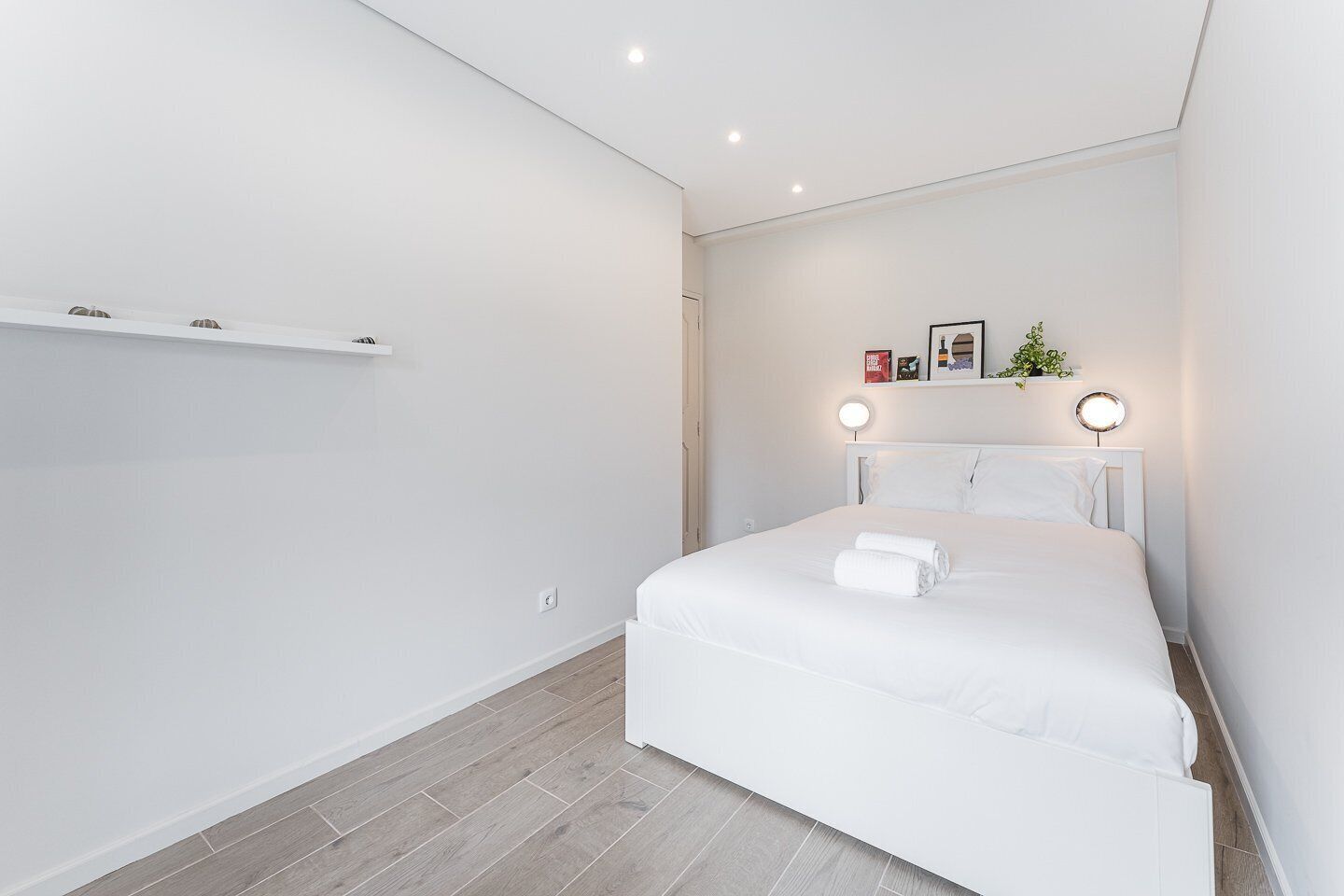 Quarto moderno com uma cama grande, paredes brancas e piso de madeira. Há duas mesas de cabeceira com luminárias e uma pequena estante sobre a cama.