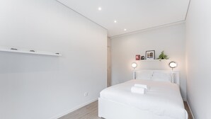 Une chambre à coucher moderne avec un grand lit, des murs blancs et un plancher en bois. On y trouve deux tables de chevet avec des lampes et une petite étagère au-dessus du lit.