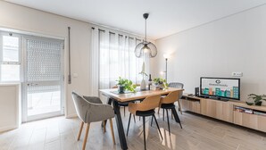 Un espace repas moderne avec une table en bois, quatre chaises, un luminaire suspendu et un téléviseur affichant le logo « GuestReady ».