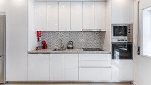 Une cuisine moderne avec des meubles blancs, un pare-feu en carrelage, un évier en acier inoxydable, une bouilloire rouge et des appareils électroménagers intégrés, notamment un four et un four à micro-ondes.