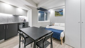 Habitación de hotel compacta con kitchenette, zona de comedor con mesa y sillas, cama con colcha azul y un ventanal con vistas al paisaje.