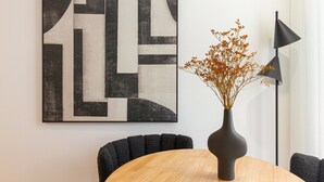 Ein moderner Essbereich mit einem runden Holztisch, einer schwarzen Vase mit Trockenblumen und einem geometrischen Wandbild.