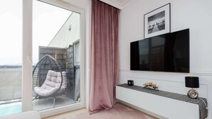 A modern living room with a large TV, a cushioned chair by a sliding glass door, and a small console table with decorative items.