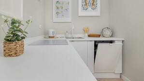 A modern kitchen with white cabinetry, a sink, and a dishwasher. There are framed botanical prints on the wall and a potted plant on the counter. - Comfortable Studio in the Center of Katowice | For 3 People (Katowice)