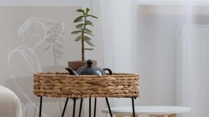 A wicker side table with a teapot and a plant, positioned next to a white chair and a small round table with wooden legs. - Comfortable Studio in the Center of Katowice | For 3 People (Katowice)